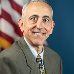 A man with white and grey hair poses for a portrait. He is wearing a tweed brown suit jacket over a light blue collared shirt with a multi-colored tie. Behind him is a blue background with an American flag over his right shoulder.