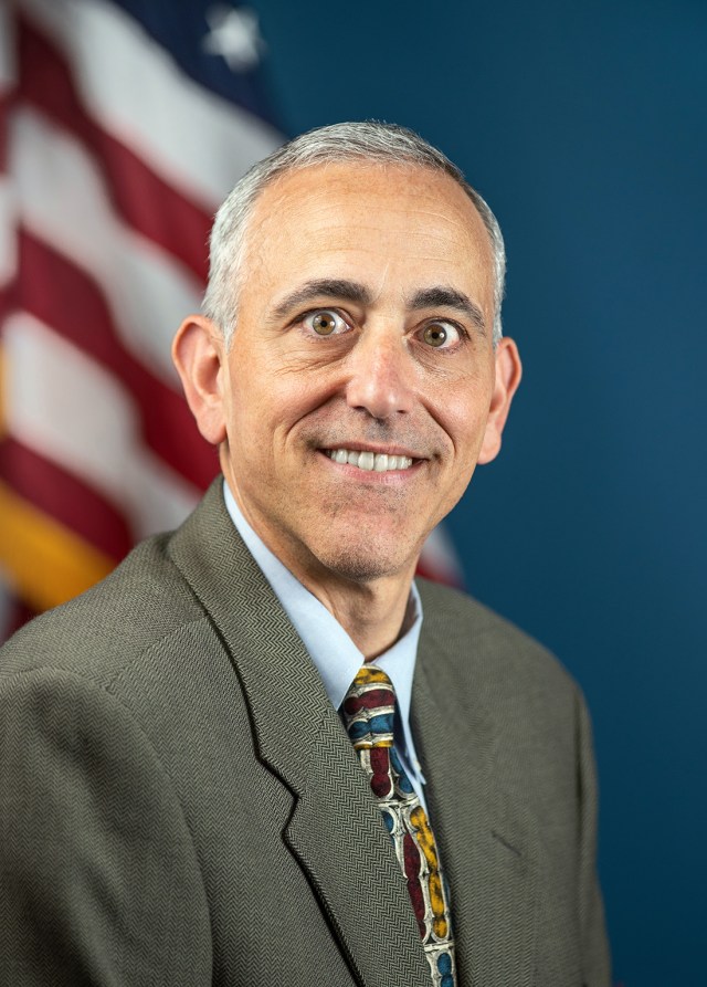 A man with white and grey hair poses for a portrait. He is wearing a tweed brown suit jacket over a light blue collared shirt with a multi-colored tie. Behind him is a blue background with an American flag over his right shoulder.