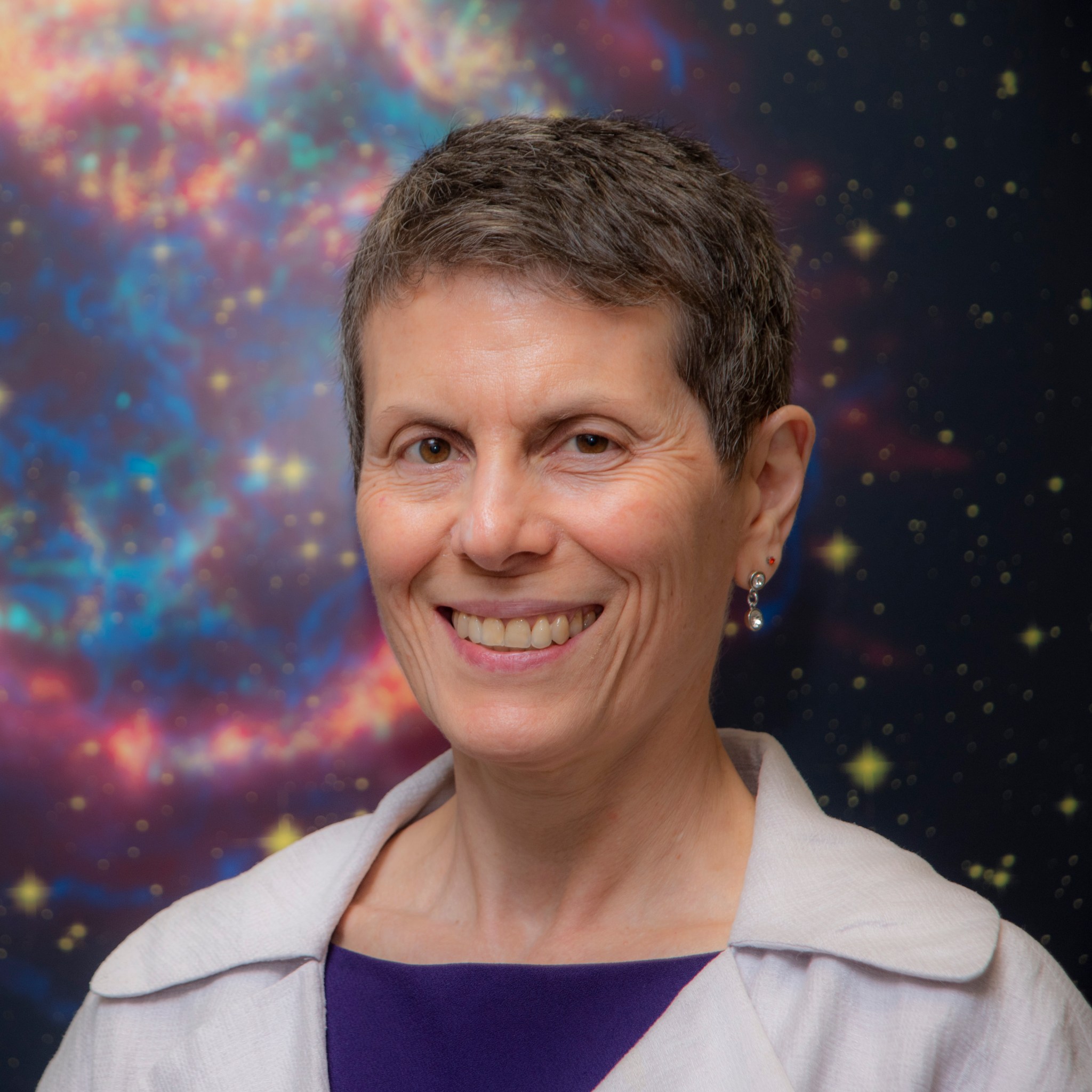 Portrait photo of Dr. Rita Sambruna smiling with a light colored jacket over a purple shirt. Behind her is a galaxy portrait.
