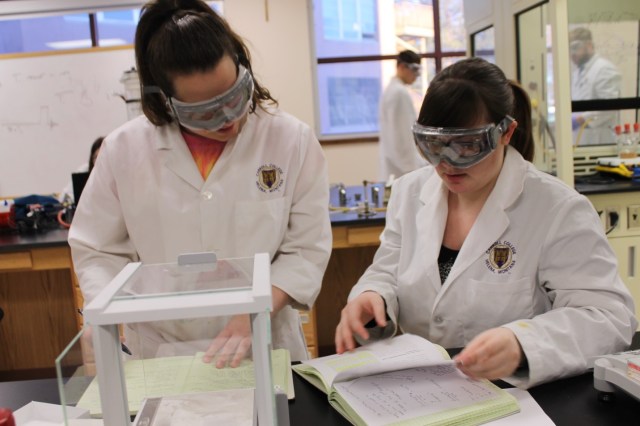 Two Students with goggles and white jackets with a college logo are working on a project.