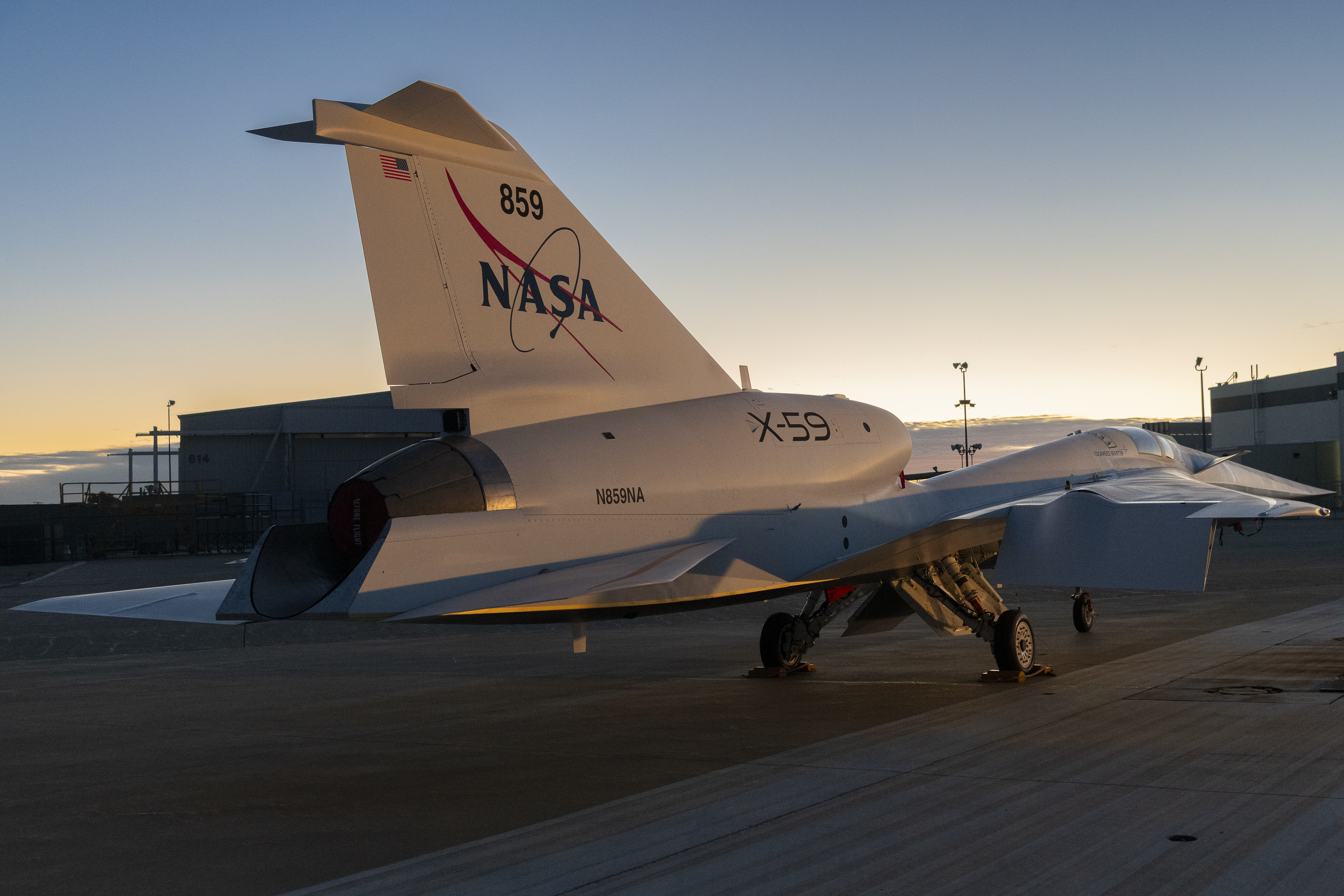 In this photo, NASA’s X-59 aircraft is shown at sunrise with its new paint scheme. The aircraft is mostly white with a blue underside and red accents on its swept back wings.
