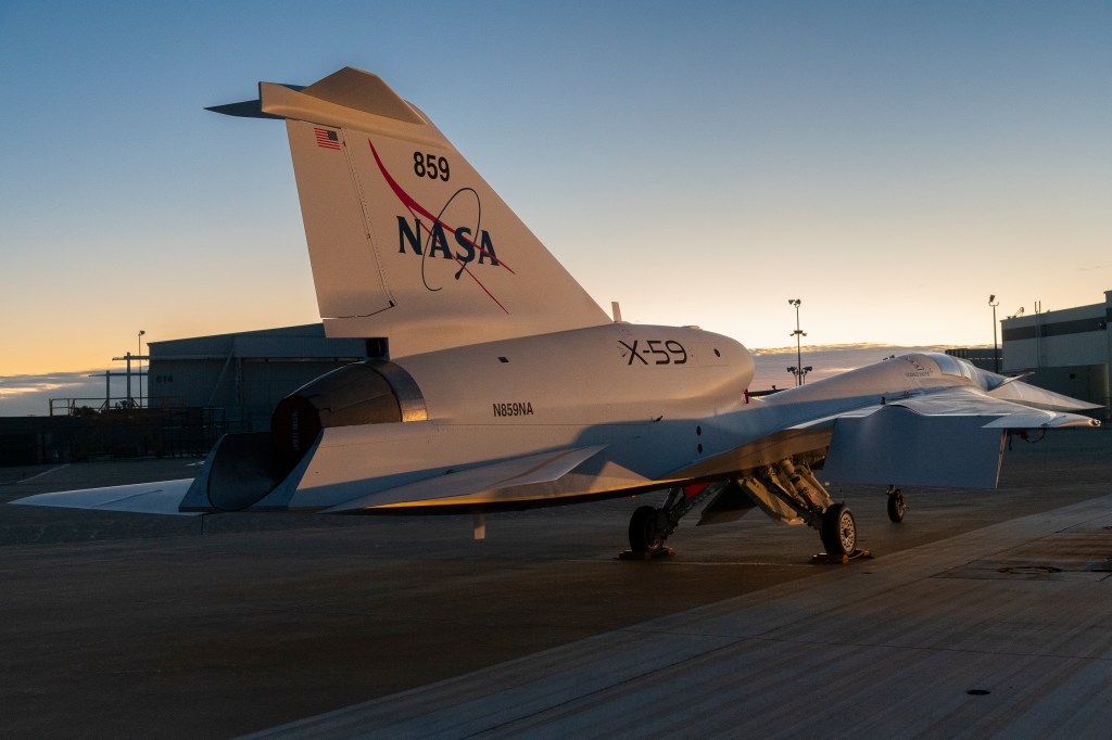 In this photo, NASA’s X-59 aircraft is shown at sunrise with its new paint scheme. The aircraft is mostly white with a blue underside and red accents on its swept back wings.