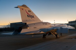 In this photo, NASA’s X-59 aircraft is shown at sunrise with its new paint scheme. The aircraft is mostly white with a blue underside and red accents on its swept back wings.