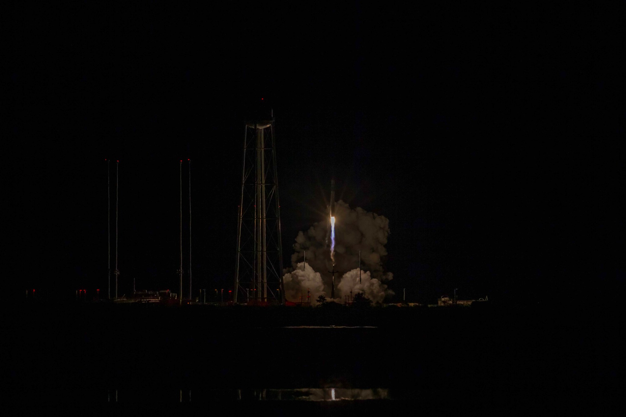 Rocket Lab's small black and white rocket launches off the pad in near pitch black. The rocket is just off the pad, surrounding by smoke. A water tower with spindly legs are to the left of the rocket, slightly lit up by the blow of the rocket. 
