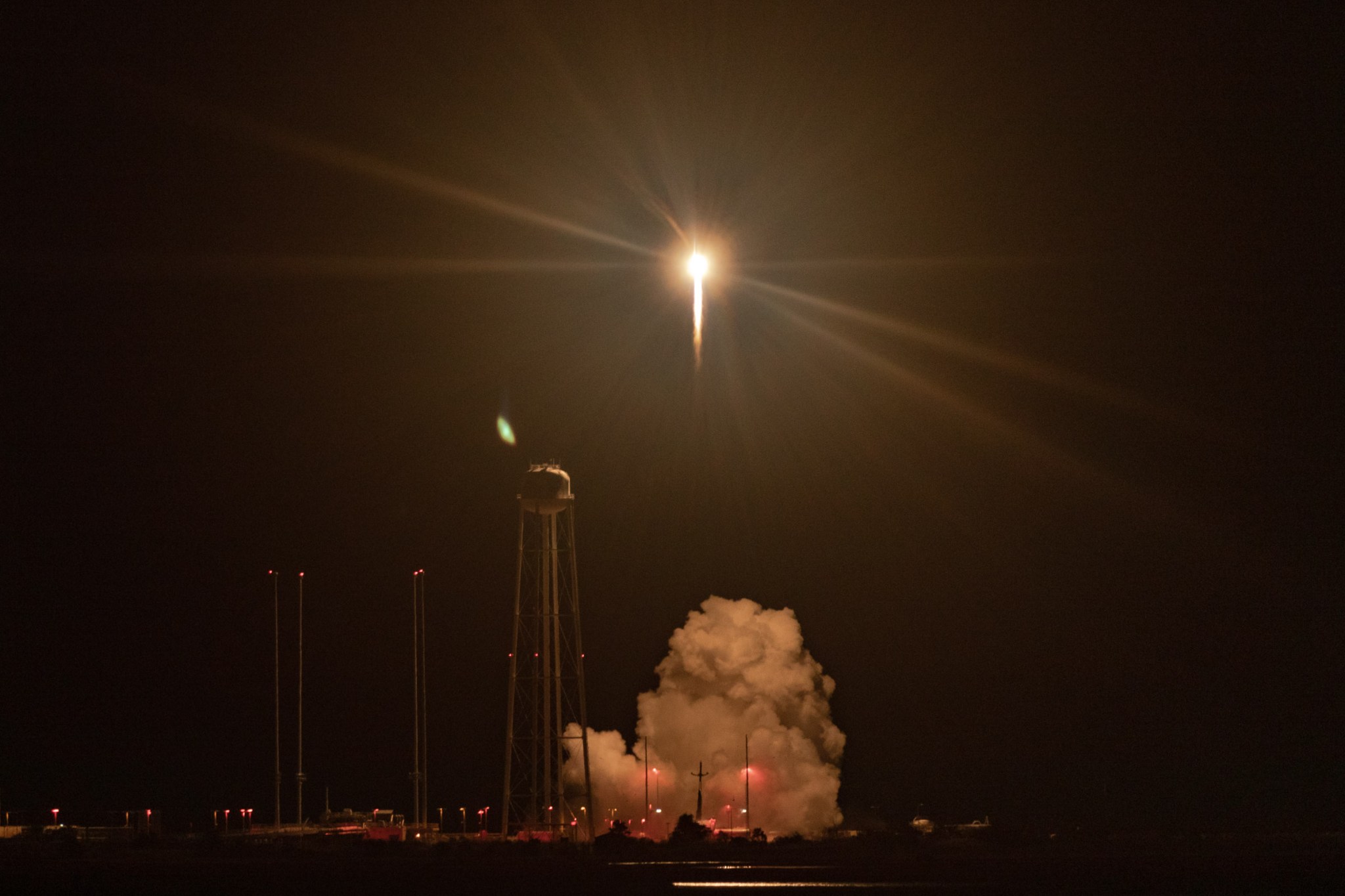 A rocket launches off the pad in near pitch black. The rocket is bright in the air with a golden starburst forming around it. A water tower with spindly legs is to left, slightly lit up by the blow of the rocket. A cloud of smoke is formed below.