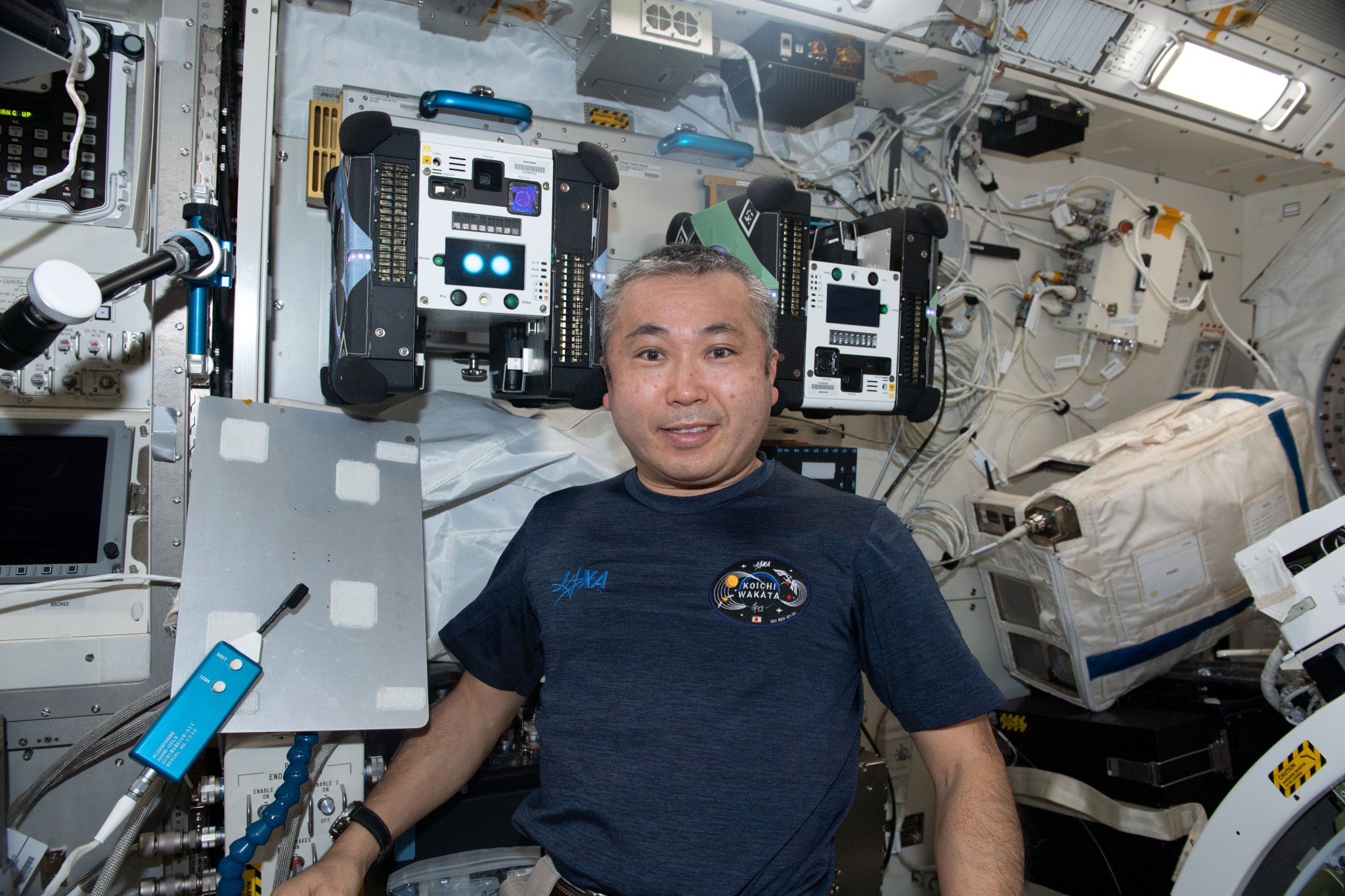 astronaut Koichi Wakata in front of the space station’s Astrobee robots floating with lots of buttons and cables behind him.