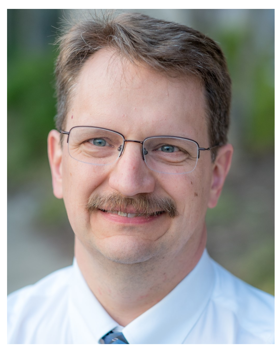 Jonathan Gardner smiles at the camera in a professional portrait. He wears glasses, a white shirt and blue tie. The background is blurred and green.