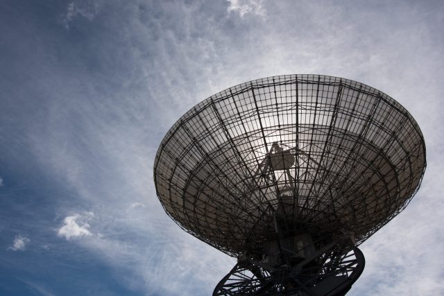 A massive Deep Space Network antenna is backlit by the Sun and a bright blue sky.