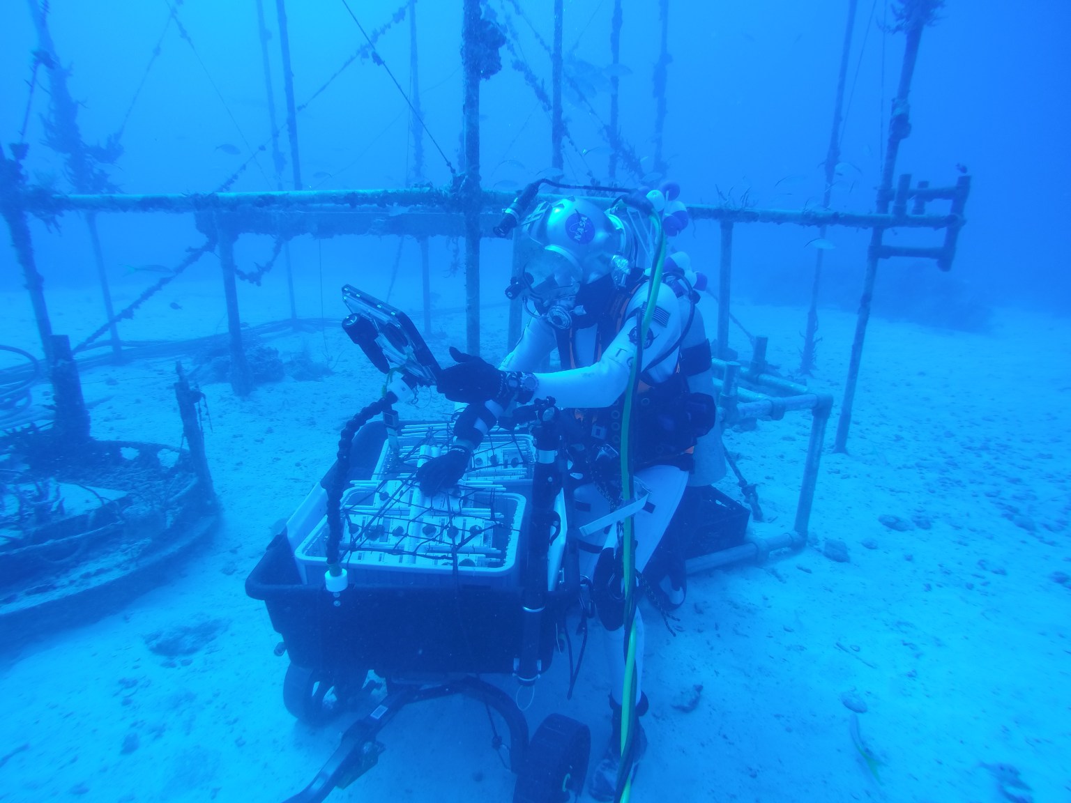Aquanaut underwater working on a small machine in front of metal rigging.