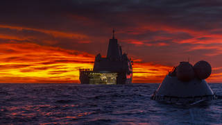 Brilliant red and yellow sunrise over the ocean with the Orion test capsule in front of the USS Murtha ship
