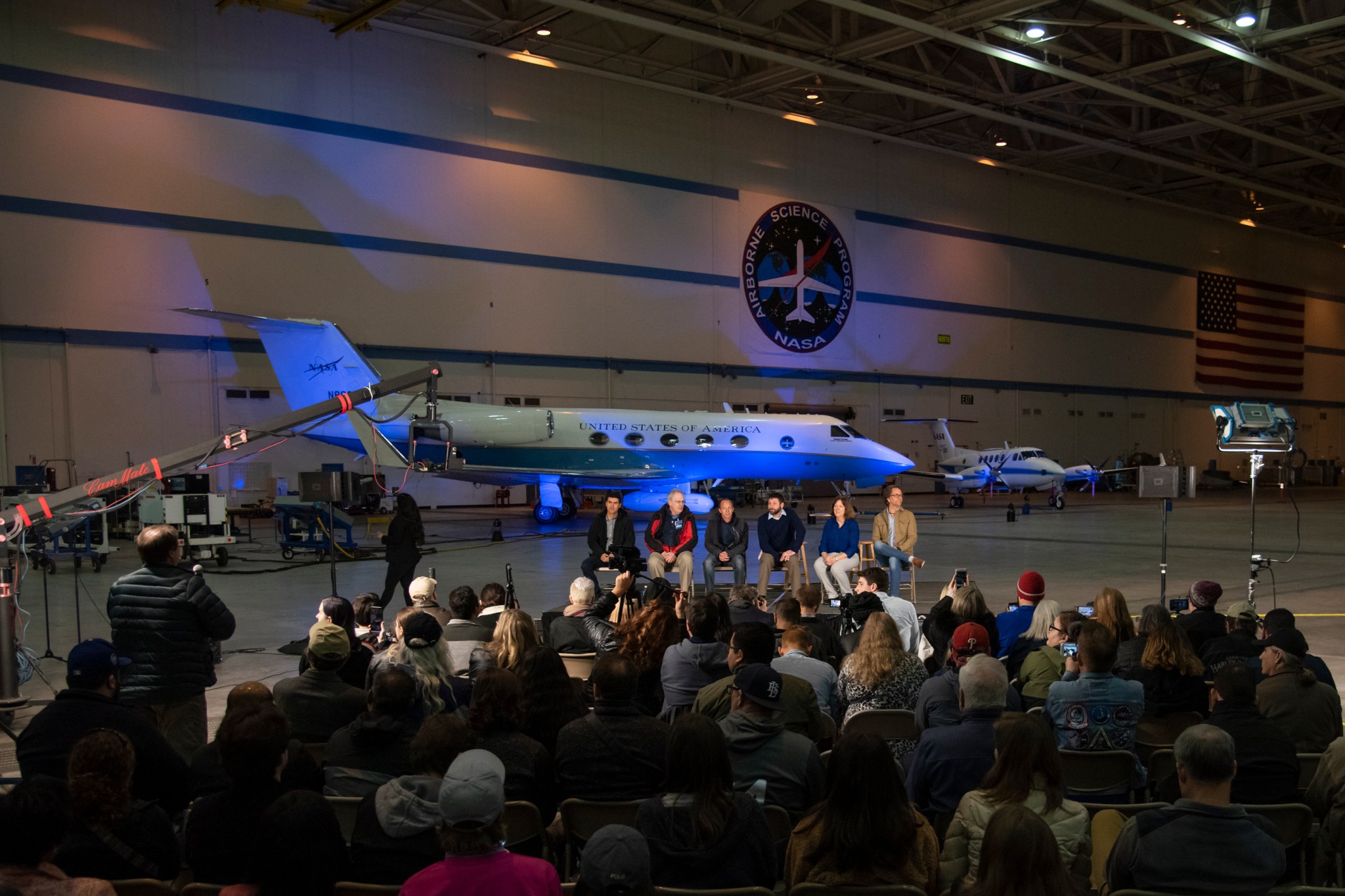 Inside a NASA Armstrong hangar, a group of six NASA airborne science representatives sit in front a large group of media. Armstrong's C-20A science aircraft is seen in the background.