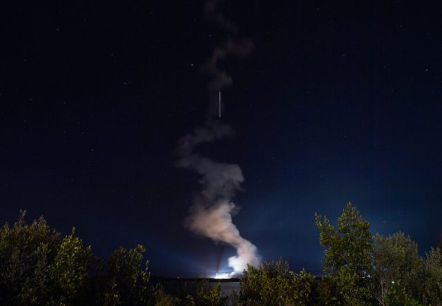 A rocket launches in the night sky with trees in the foreground.