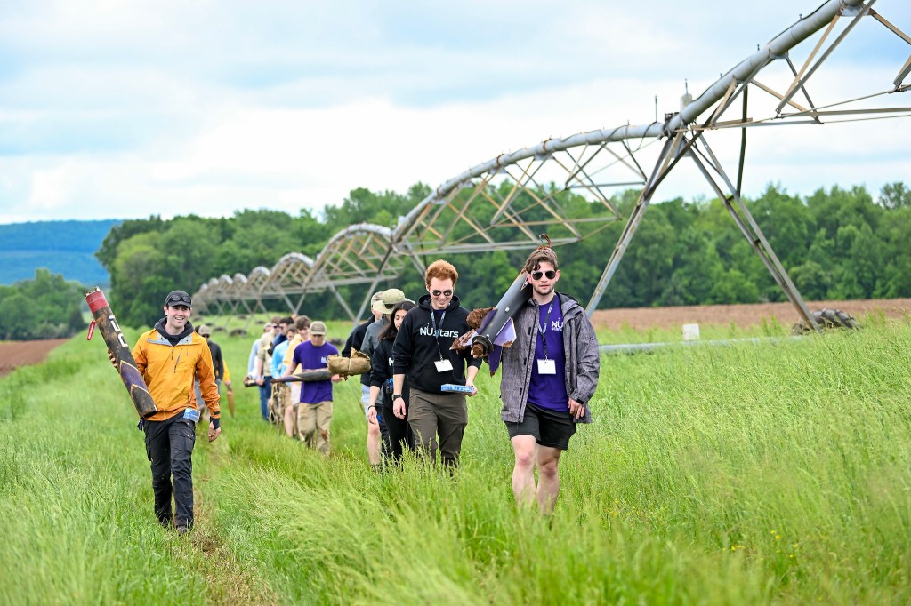 Students walk in a straight line in a field, carrying parts of a rocket after competing in the 2025 Student Launch Challenge