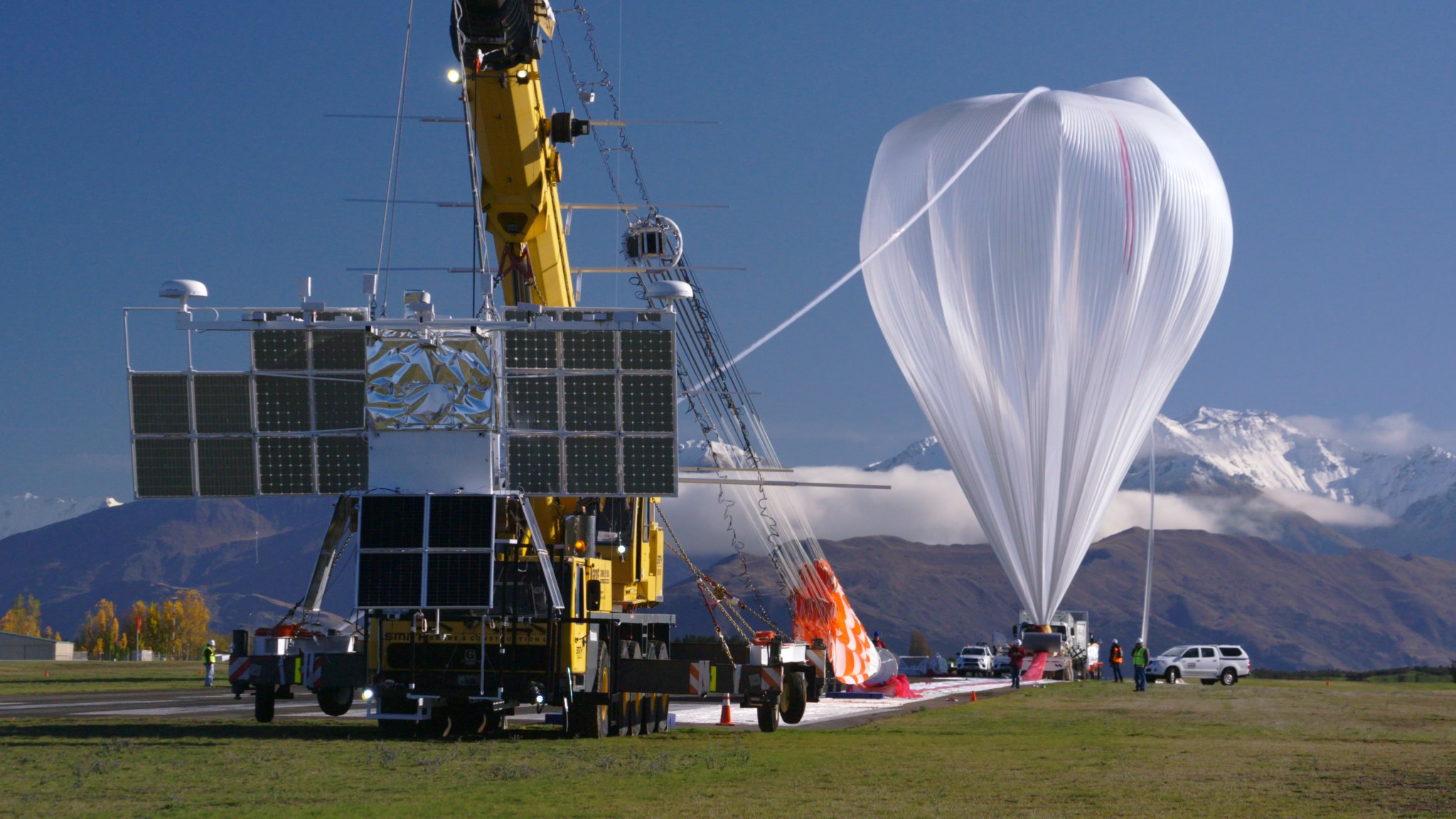A crane is leading a scientific balloon before launch. The balloon is to the right, and appears an a plastic, upside down teardrop. A tube attached to the top of the balloon leads down to the ground where a number of personnel are holding it. To the left, a crane holding a large payload structure with many solar panels is attached to the end of the balloon.