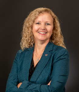 Christa Peters-Lidard smiles at the camera in an official portrait. She has long blonde hair and wears a teal suit jacket, black blouse, and necklace. The background is brown.