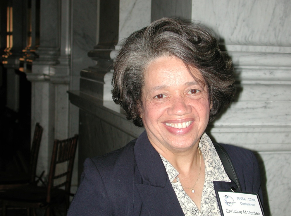 Christine Mann Darden smiles at the camera in a brightly lit snapshot. She has short gray hair and wears a navy suit jacket, floral blouse, and nametag. The background is marble columns in dark shadow.
