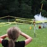 A young girl with a long blond ponytail and black T-shirt faces away from the camera, watching a small model rocket lift off from its launch pad. The rocket is behind yellow caution tape, and a thin stream of white smoke billows out from its end into a small white cloud underneath as it shoots diagonally up toward the upper right corner of the image. The grass between the child and the rocket is green, and the background is dense, dark green trees.