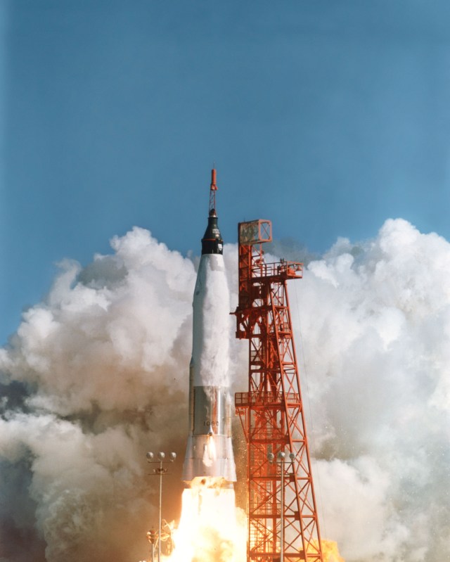 A vintage photo of a rocket lifting off from the launch pad. The rocket is white with a black, conical metal capsule at the top and a red "escape tower," or tiny red rocket attached to the end of the capsule. Bright white fire shoots out of the rocket's end, and white smoke billows around the bright red frame of the launch tower beside the rocket. The sky is bright blue.
