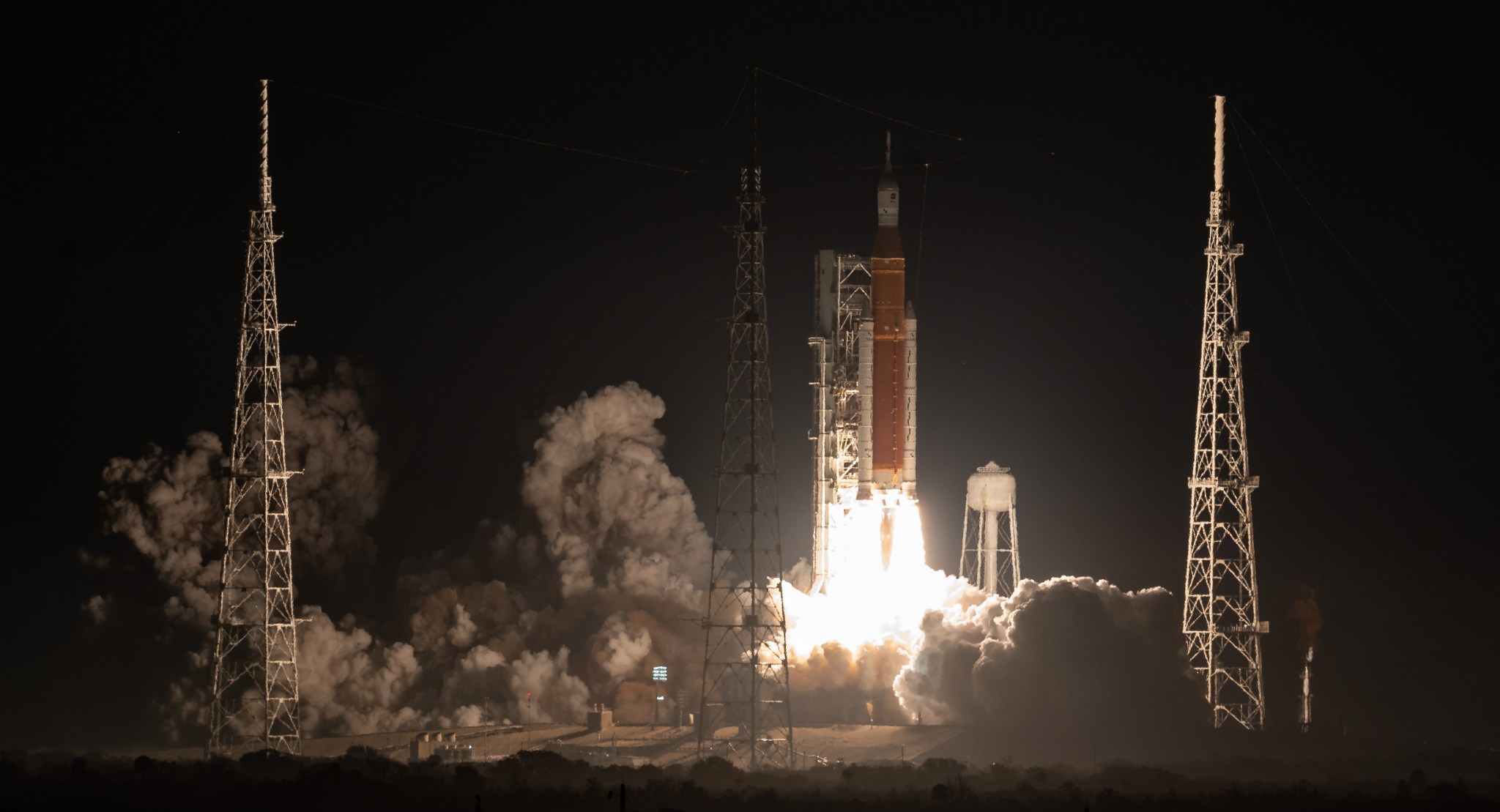 The Artemis I Space Launch System rocket carrying Orion lifts off from its launch pad, lighting up the night sky with its fiery back end. Smoke billows out from the side.