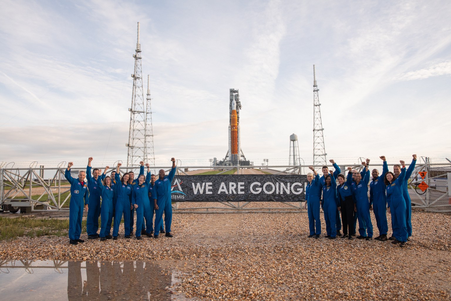 Eighteen astronauts and astronaut candidates in blue flight suits each raise a fist as they pose for a photo outside the gate of Launch Pad 39B.