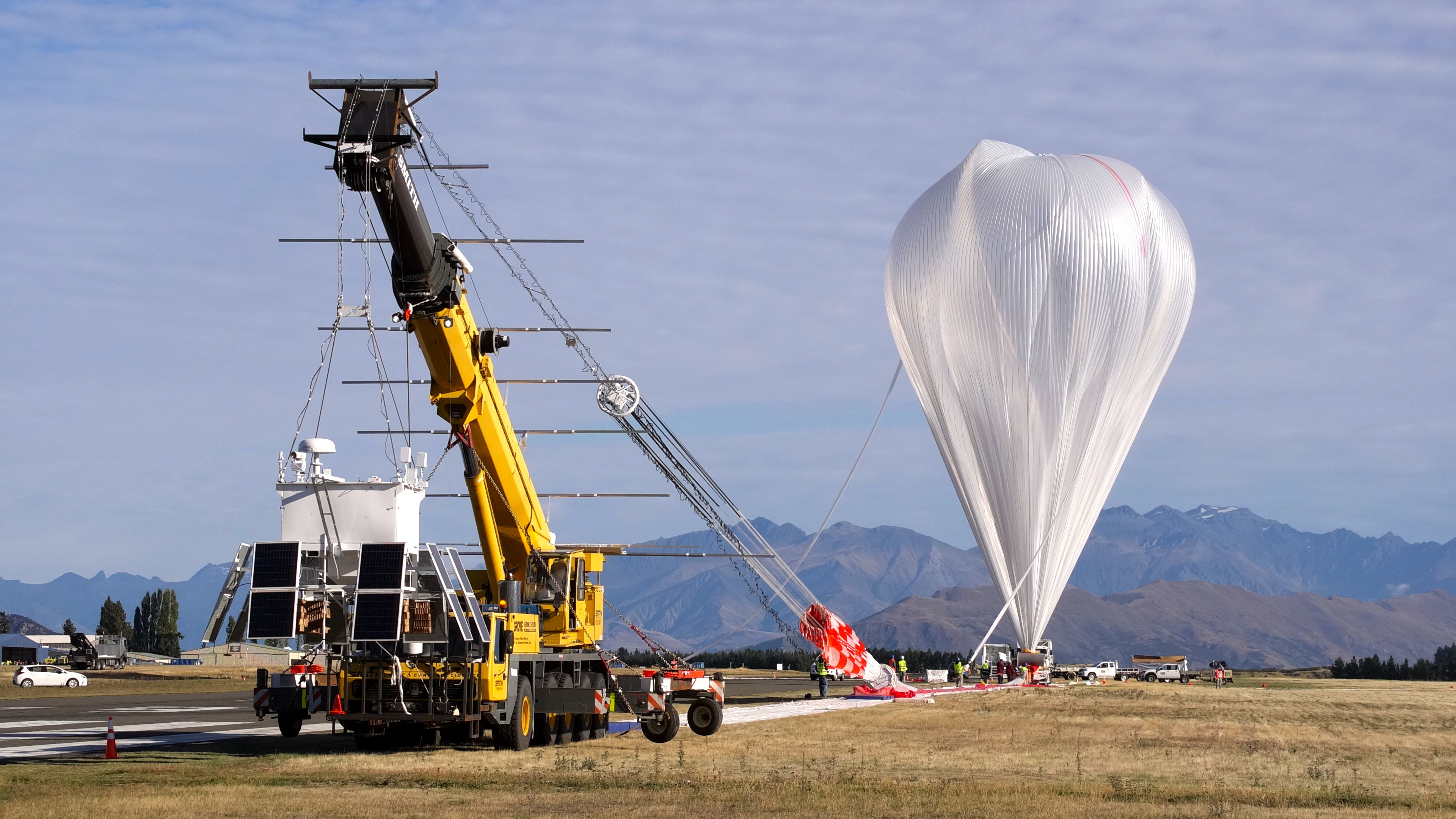A crane is leading a scientific balloon before launch. The balloon is to the right, and appears an a plastic, upside down teardrop. A tube attached...