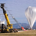 A crane is leading a scientific balloon before launch. The balloon is to the right, and appears an a plastic, upside down teardrop. A tube attached to the top of the balloon leads down to the ground where a number of personnel are holding it. To the left, a crane holding a large payload structure with many solar panels is attached to the end of the balloon.