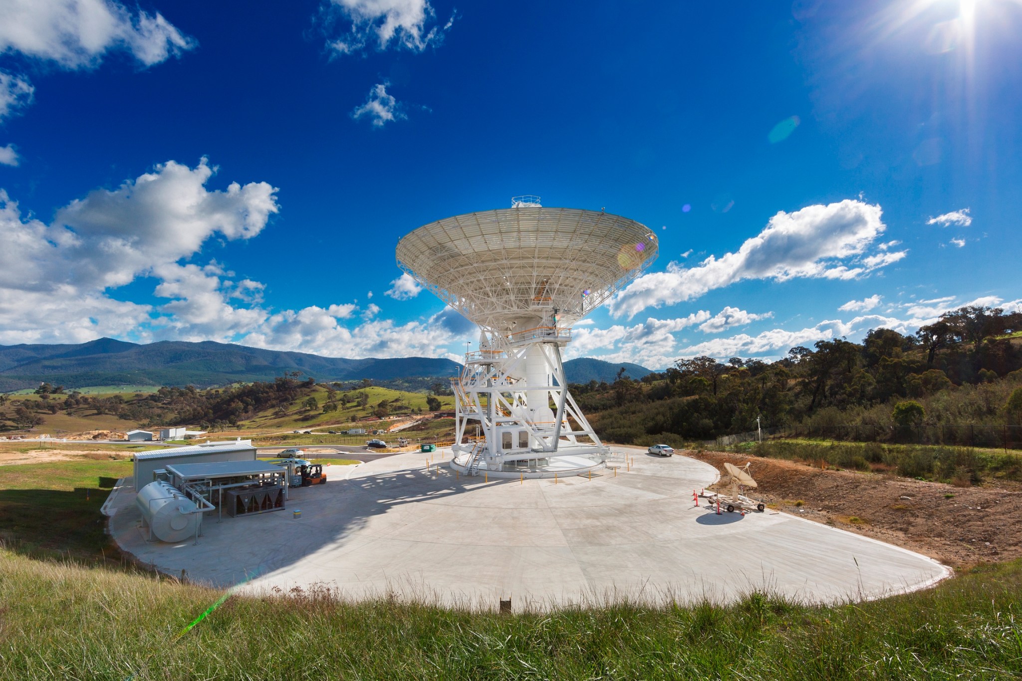 A 111-foot (34-meter) antenna points toward a bright blue sky. Mountains and several clouds can be seen in the background.