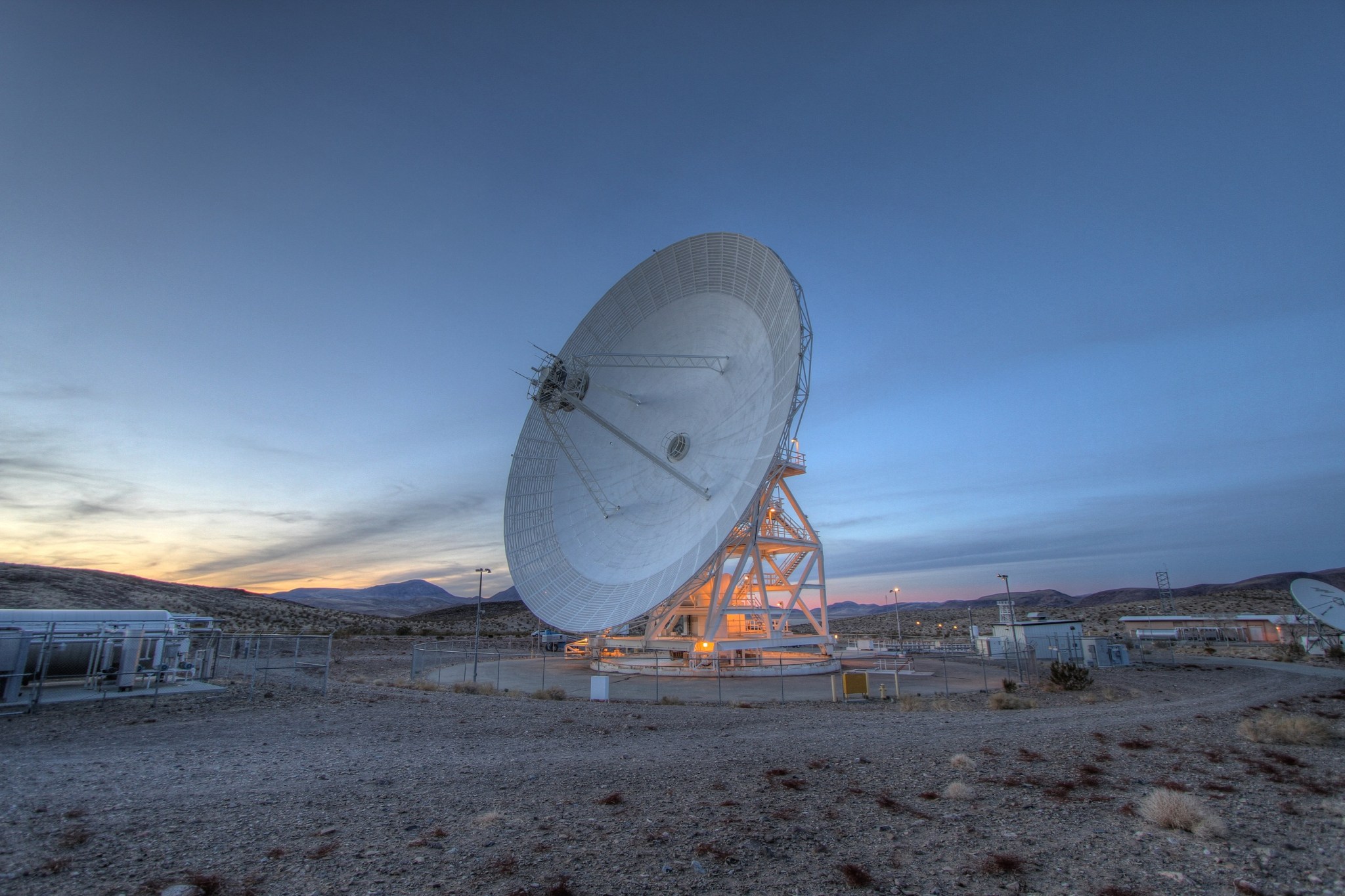 An antenna faces left, surrounded by a rocky landscape, it casts an orange glow in the darkening sky. The Sun peaks behind a mountain range on the left as it sets.