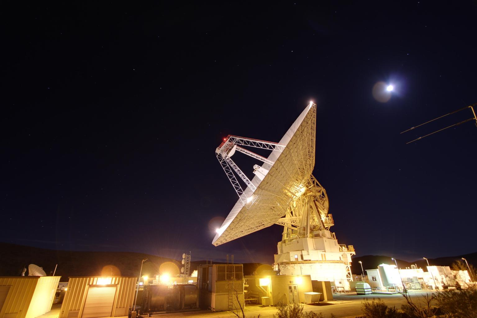 A 70-meter antenna lights up the night sky at the Goldstone Deep Space Communications Complex near Barstow, California. The Moon brightly shines behind the giant antenna.