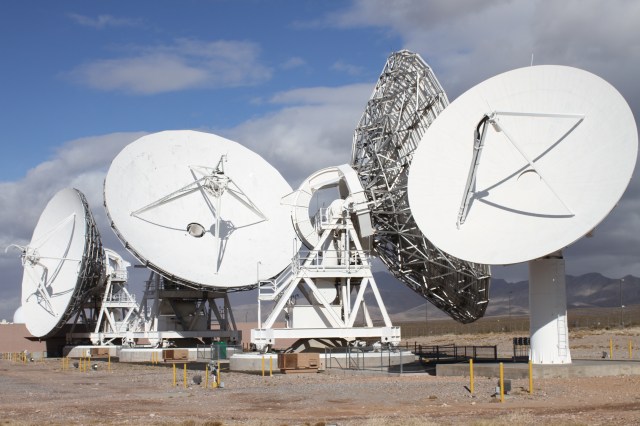 Four white antennas face the Sun, surrounded by desert and a mountainous landscape.