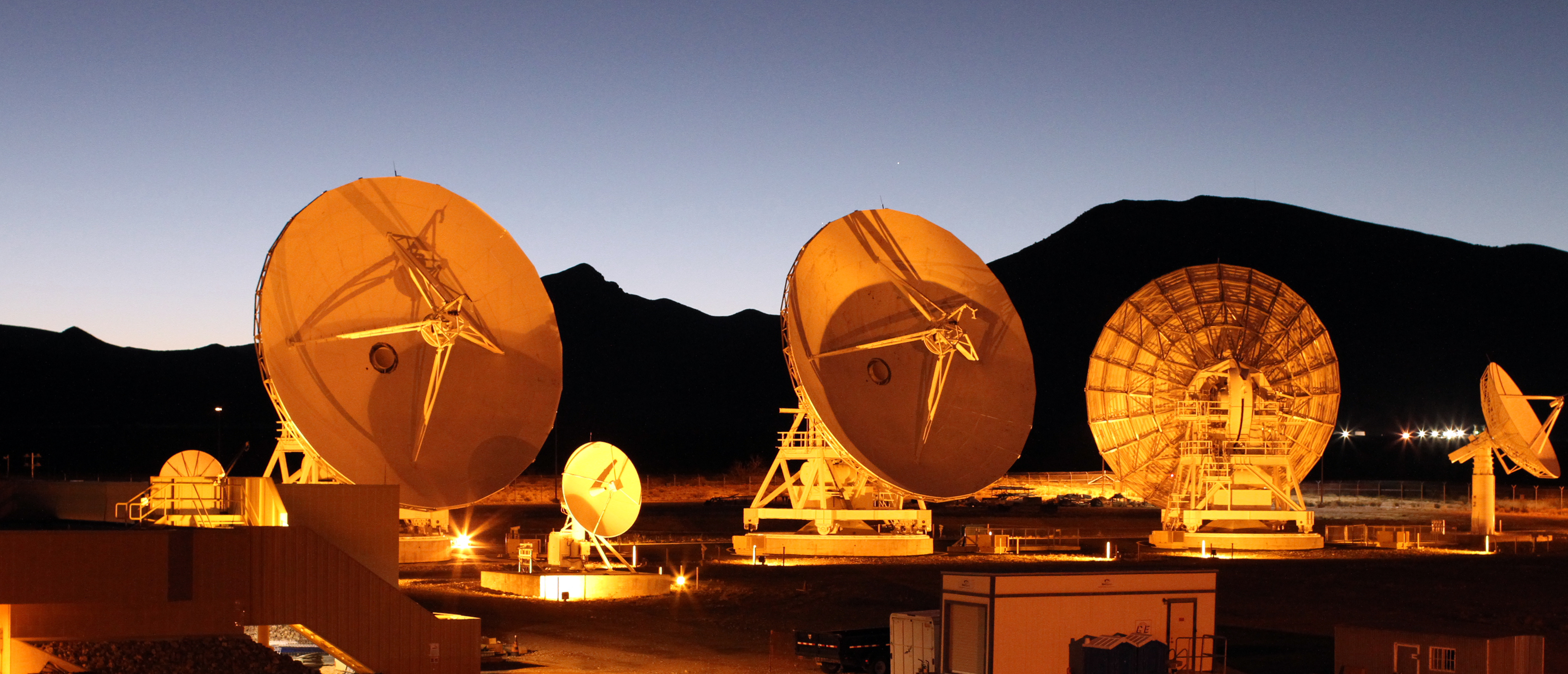 Several antennas point in varying directions. The Sun is rising behind a mountain range in the background, casting an orange glow across the antenn...