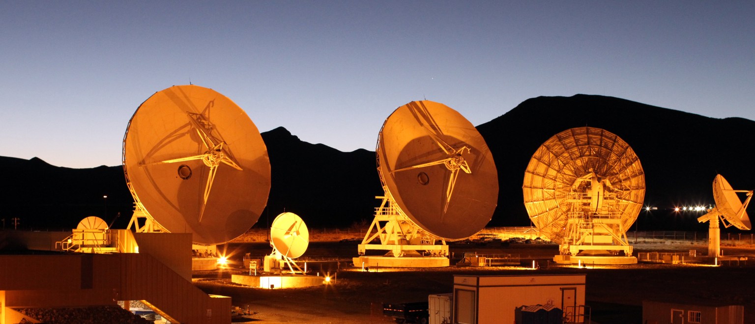Several antennas point in varying directions. The Sun is rising behind a mountain range in the background, casting an orange glow across the antennas.