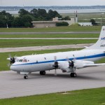 The P-3 Aircraft is a white plane with a blue strip going down the middle of the plane. An older version of a NASA logo is on the tail. The words NASA are blue crossed by a red, V-shaped swoosh. The plane is on a runway with its propellers spinning before takeoff.