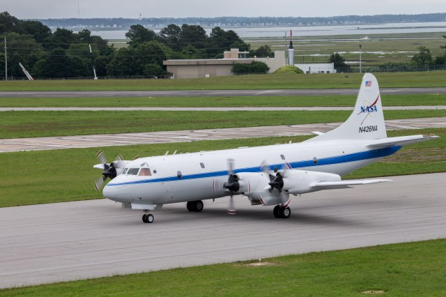 The P-3 Aircraft is a white plane with a blue strip going down the middle of the plane. An older version of a NASA logo is on the tail. The words NASA are blue crossed by a red, V-shaped swoosh. The plane is on a runway with its propellers spinning before takeoff.