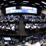 A large control room with wide screen of monitors with different views. There are three rows of desks, all with monitors at each station. There is another row of desks with monitors toward the bottom of the image. People are stationed throughout the control center.