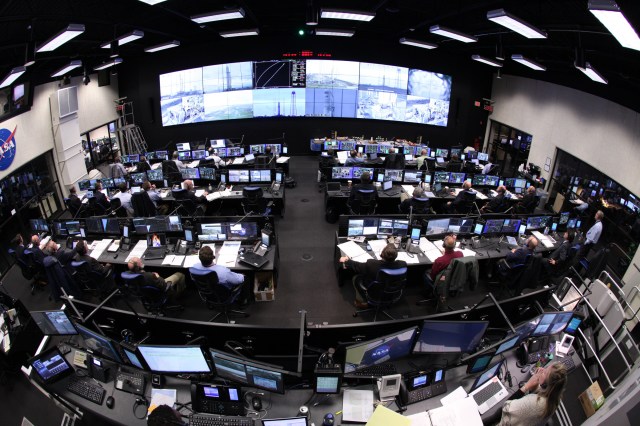 A large control room with wide screen of monitors with different views. There are three rows of desks, all with monitors at each station. There is another row of desks with monitors toward the bottom of the image. People are stationed throughout the control center.