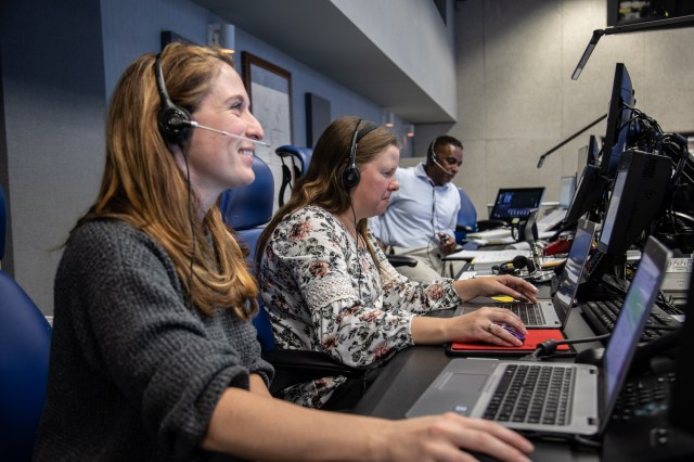 A photo of a group of three people taken from the side sitting at a long desk. The closest person is a women with long, light-brown hair wearing a grey sweatshirt and a headset and smiling. To her left is another women with long, light-brown hair with a floral shirt and a headset working at a laptop. To her left is a black man with a light-blue button down dress shirt and headset looking down at paperwork.