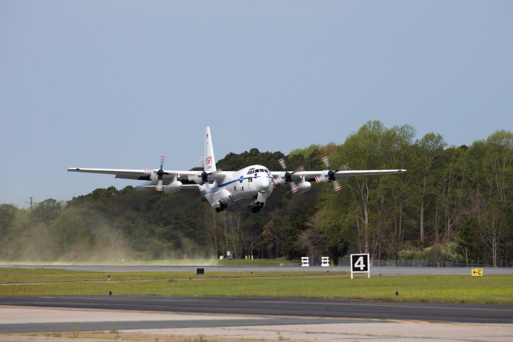 A white plane, with a blue strip down the side, blue NASA meatball logo in the front, and red NASA worm logo on the tail fin takes off from a runway, kicking up a dust cloud of bright green pollen just as it takes off. A green treeline is in the background and a concrete portion of the runway in the foreground, with a grass strip separating the runway from the concrete.