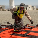 A man kneels down to work on a multi-rotor drone sitting on the pavement.