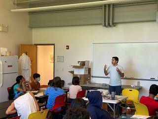 Peyman Abbaszadeh, wearing a white tee and jeans, gestures while speaking at the front of a classroom of children. The room has lab coats, a fridge and a large whiteboard. Children sit in colorful chairs and listen.