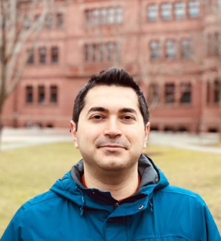 Peyman Abbaszadeh smiles slightly at the camera in an informal portrait. He has short dark hair, wears a blue winter jacket, and stands in front of an old, red brick building and yellow-green grass.