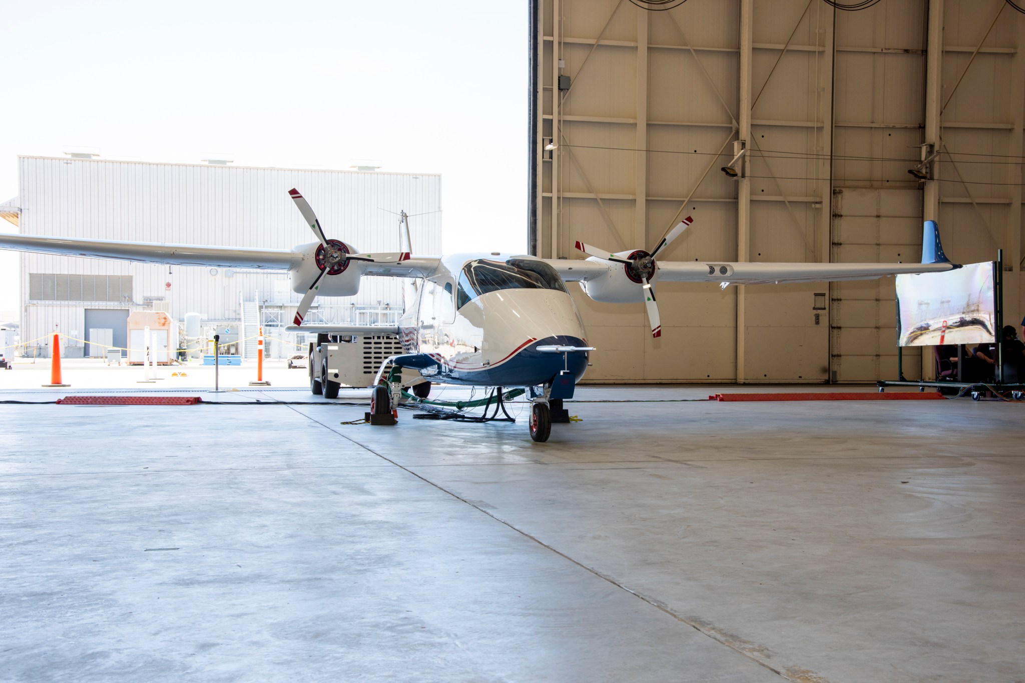 White and blue aircraft sits on concrete floor of airplane hangar attached to electric cables.