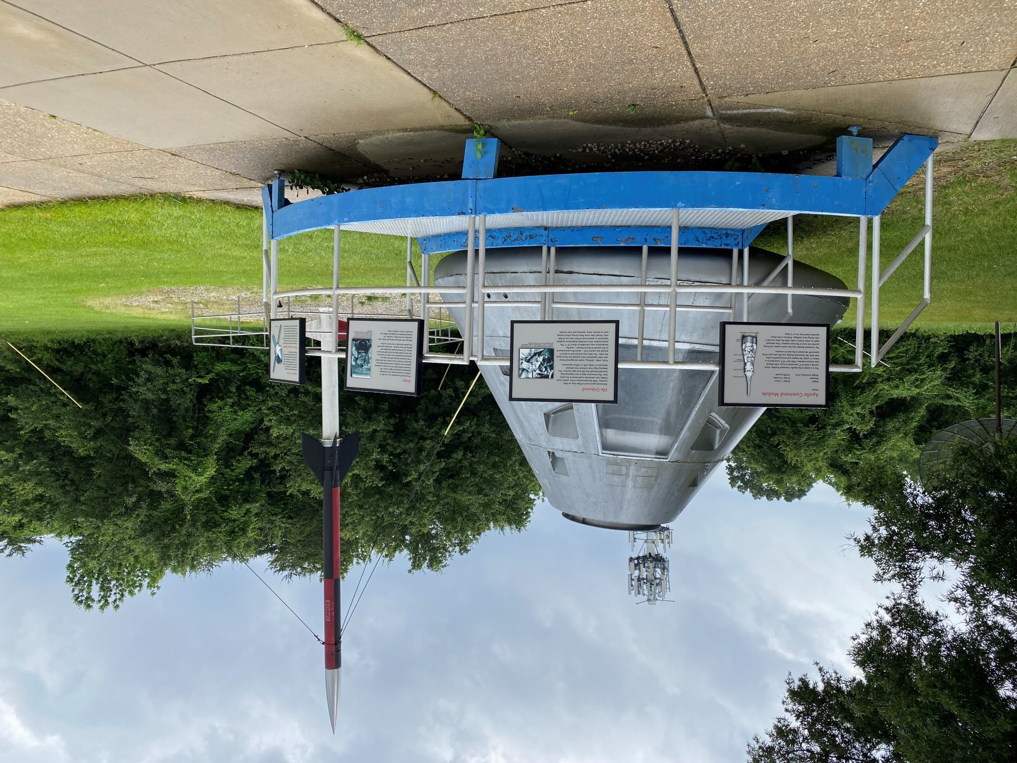 A conical Apollo-era capsule sits outdoors at the edge of a paved area, surrounded by a slightly elevated blue catwalk. The capsule is silver and leans slightly to the right. Placards with text and graphics are mounted on the catwalk's railing. Grass and green trees are further back in the background, as well as a small black-and-red rocket with a white nose cone.