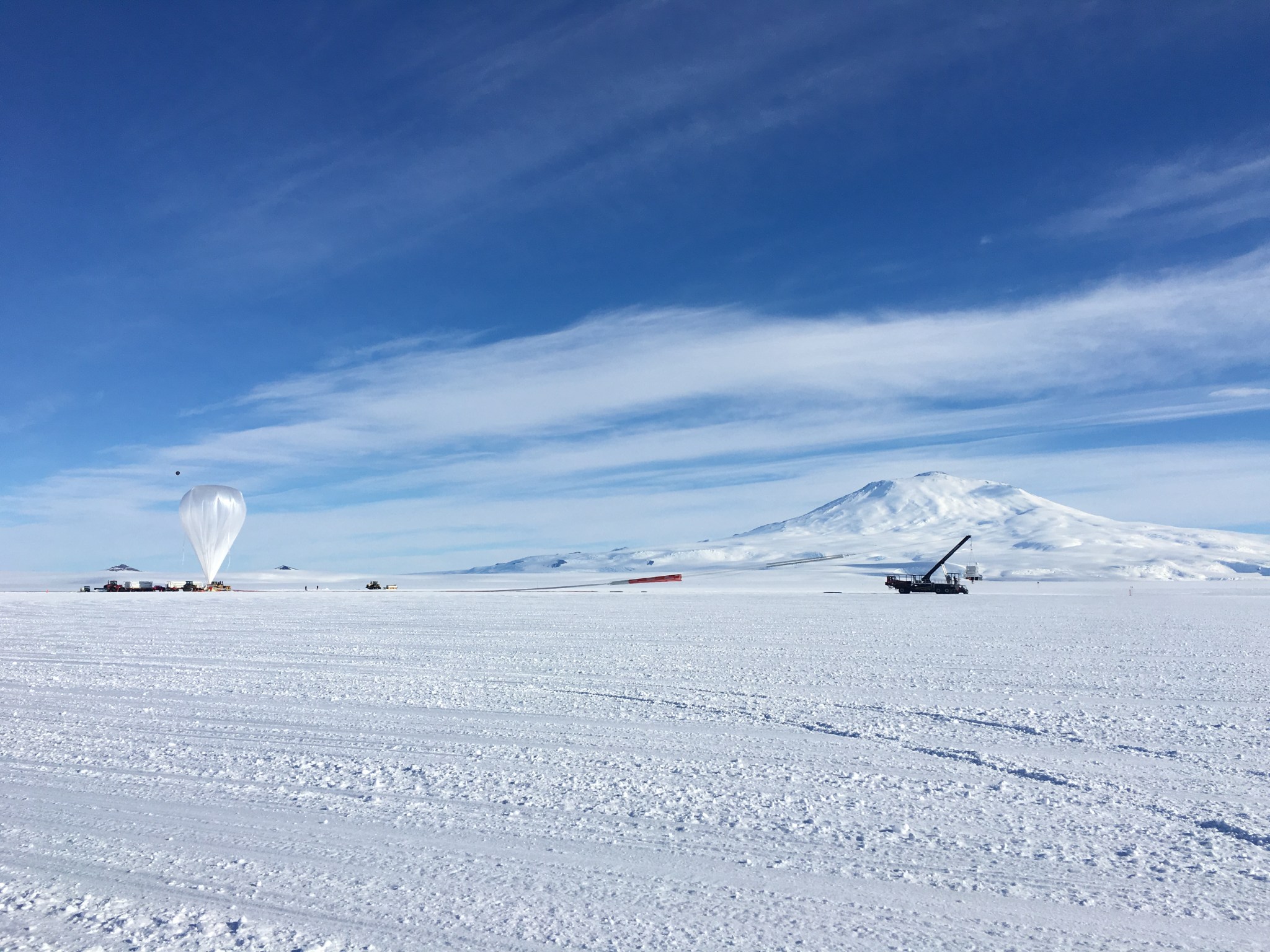 A scientific balloon being prepared for launch on a snowy landscape. The balloon is a clear plastic in the shape of an upside down tear drop. To the right is a large crane holding a square payload. The ground is completely covered in snow, and a snow-covered mountain is seen in the distance. The sky is bright blue with wispy clouds.