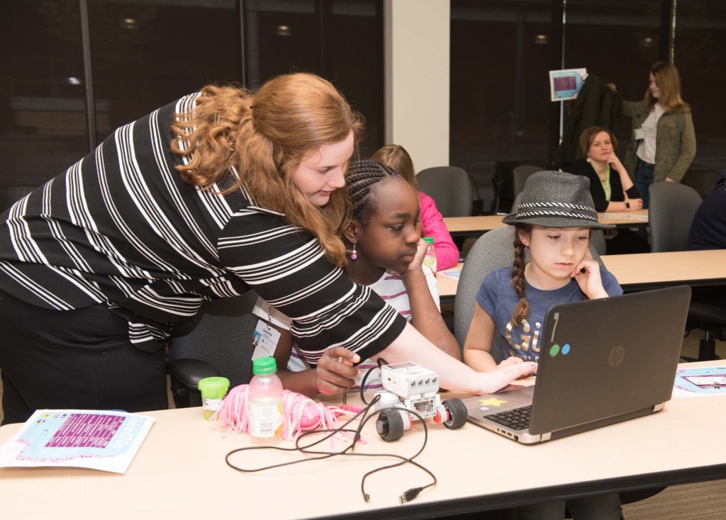 A mentor leans over the computer of two students who watch intently.
