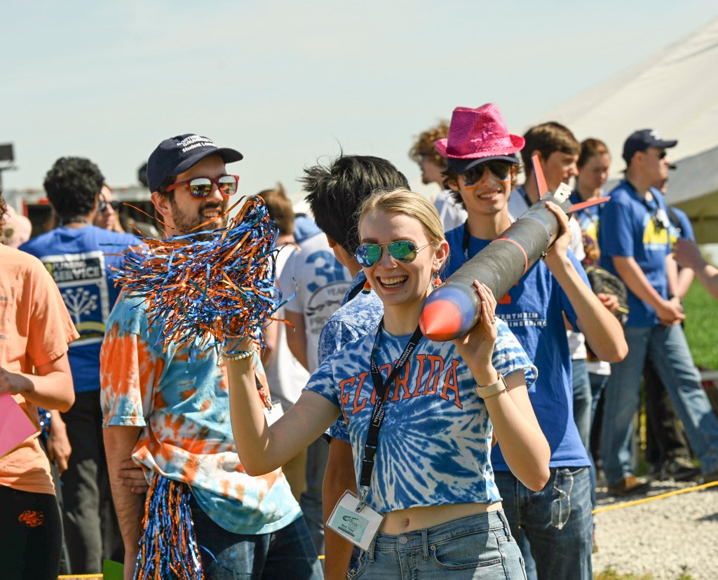 Student Launch participant shakes a pom pom and smiles while helping to carry her team's rocket.