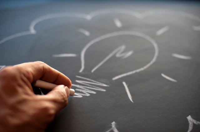 Left hand drawing a lightbulb in white chalk on a blackboard.