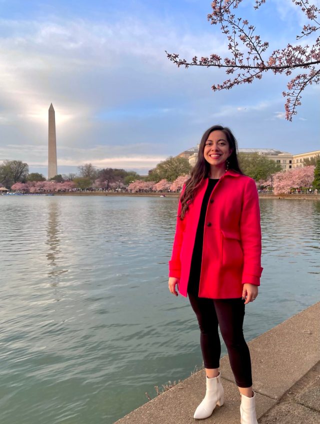 Corrine stands to the right side of the image wearing a bright red colored coat. Just above her is a branch of a cherry blossom tree with pink flowers blooming on it. To the left of Corrine is a blue green colored shimmering lake, and in the background of the image, the Washington Monument stands tall with the sun shining directly behind it. The monument is reflecting down onto the lake as well.