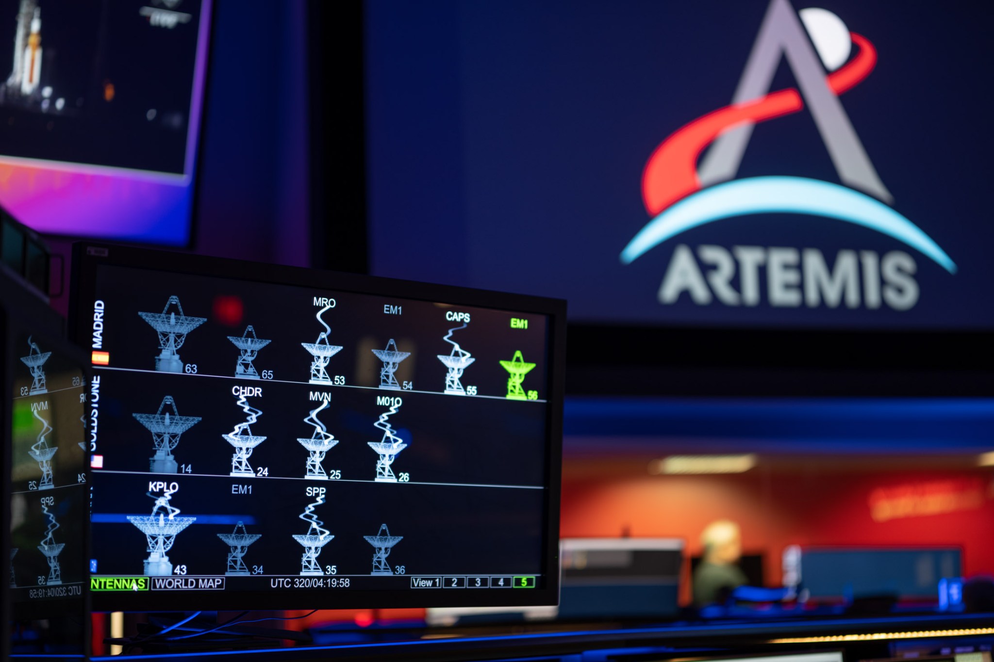 The DSN Now tool is displayed on a screen in the foreground—antenna images show real-time data provided by the Deep Space Network ground stations. The Artemis logo is seen in the background on a large screen.