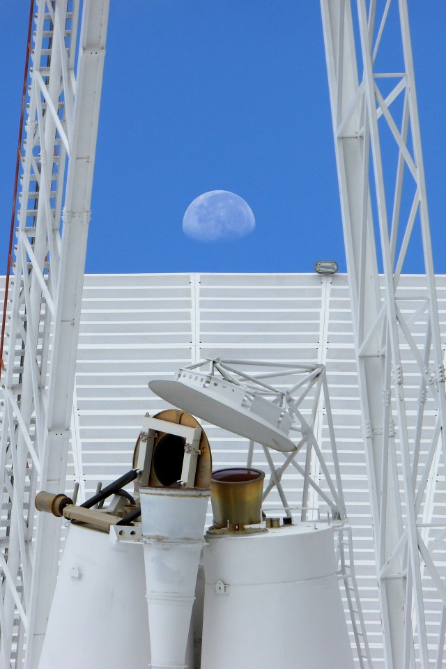 A close-up photo of a Deep Space Network antenna. Behind the antenna, the Moon can be seen clearly on a bright blue sky.